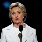 Clinton vs Trump: The Child Detention Debate Heats Up A woman in a white suit speaking at a podium during a conference