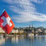 Swiss Population Cap Could Rattle EU Ties Swiss flag waving in front of Lake Lucerne with a city skyline in the background