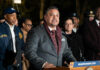 A man in a suit speaking at a podium with city officials in the background