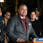 A man in a suit speaking at a podium with city officials in the background