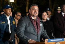 A man in a suit speaking at a podium with city officials in the background
