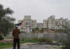 Person holding a Palestinian flag in front of buildings and a barrier