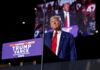 A man in a suit stands at a podium with a microphone during a political rally