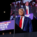 A man in a suit stands at a podium with a microphone during a political rally